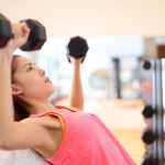 Gym woman strength training lifting dumbbell weights in shoulder press exercise. Female fitness girl exercising indoor in fitness center. Beautiful fit mixed race Asian Caucasian model training.