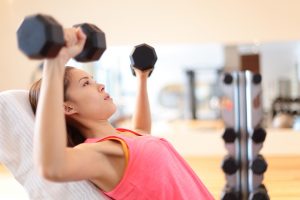 Gym woman strength training lifting dumbbell weights in shoulder press exercise. Female fitness girl exercising indoor in fitness center. Beautiful fit mixed race Asian Caucasian model training.